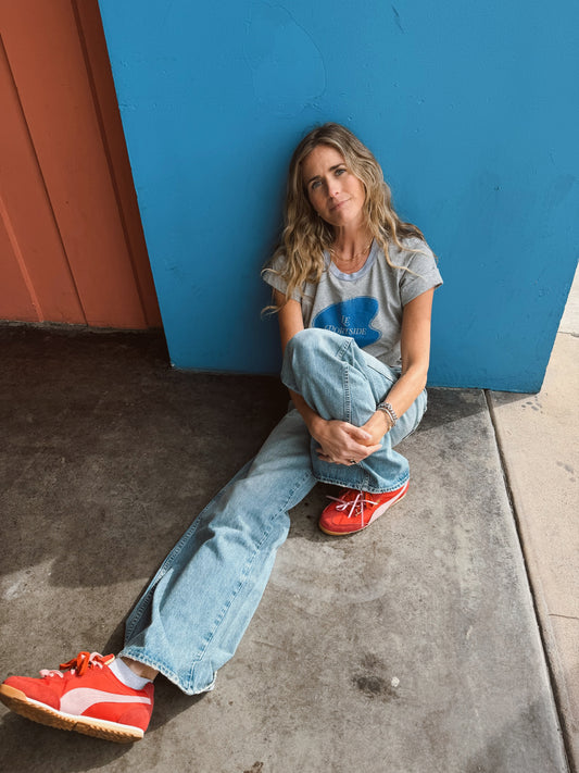 Woman wearing a heather grey rolled sleeve tee sits by a blue wall, gazing thoughtfully at the camera.