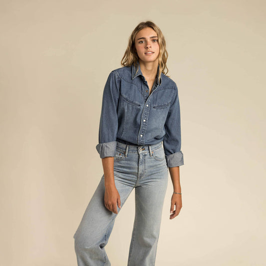 Woman wears a white pearl snap button-up shirt and light blue denim jeans, standing before a plain beige backdrop.