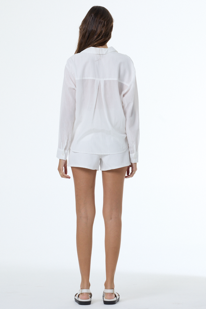 Woman facing away in a bone-colored Traveler top, white shorts, and sandals, standing against a plain background.