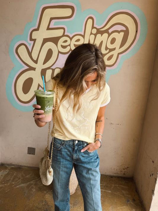 Woman in a buttercream short-sleeve cotton ringer tee stands in front of a mural with bold lettering.