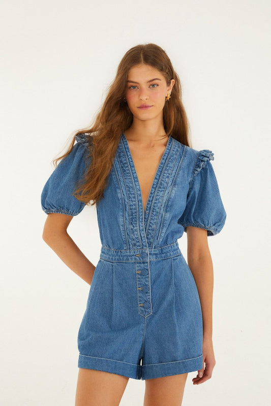 Woman wearing a blue denim ruffle romper, shown from the front against a plain white background.