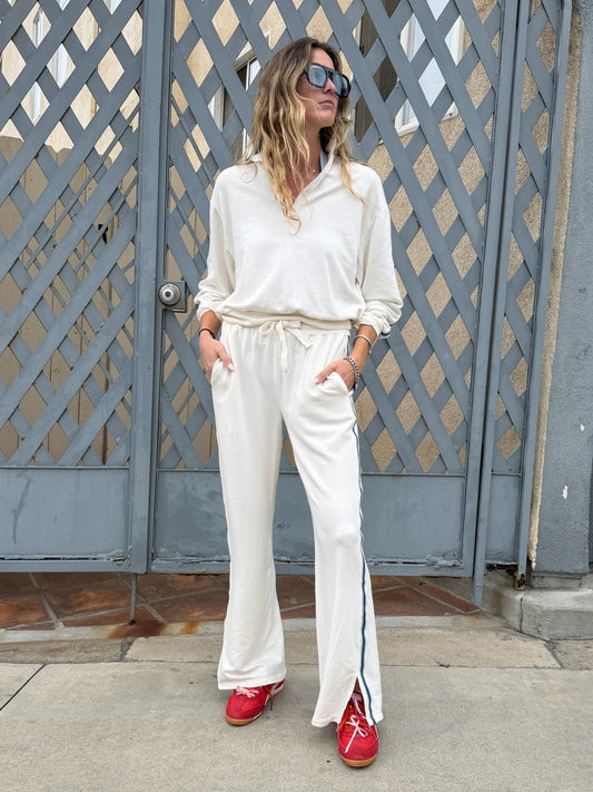 Woman in cream track pants and red shoes stands by a gray gate, facing sideways.