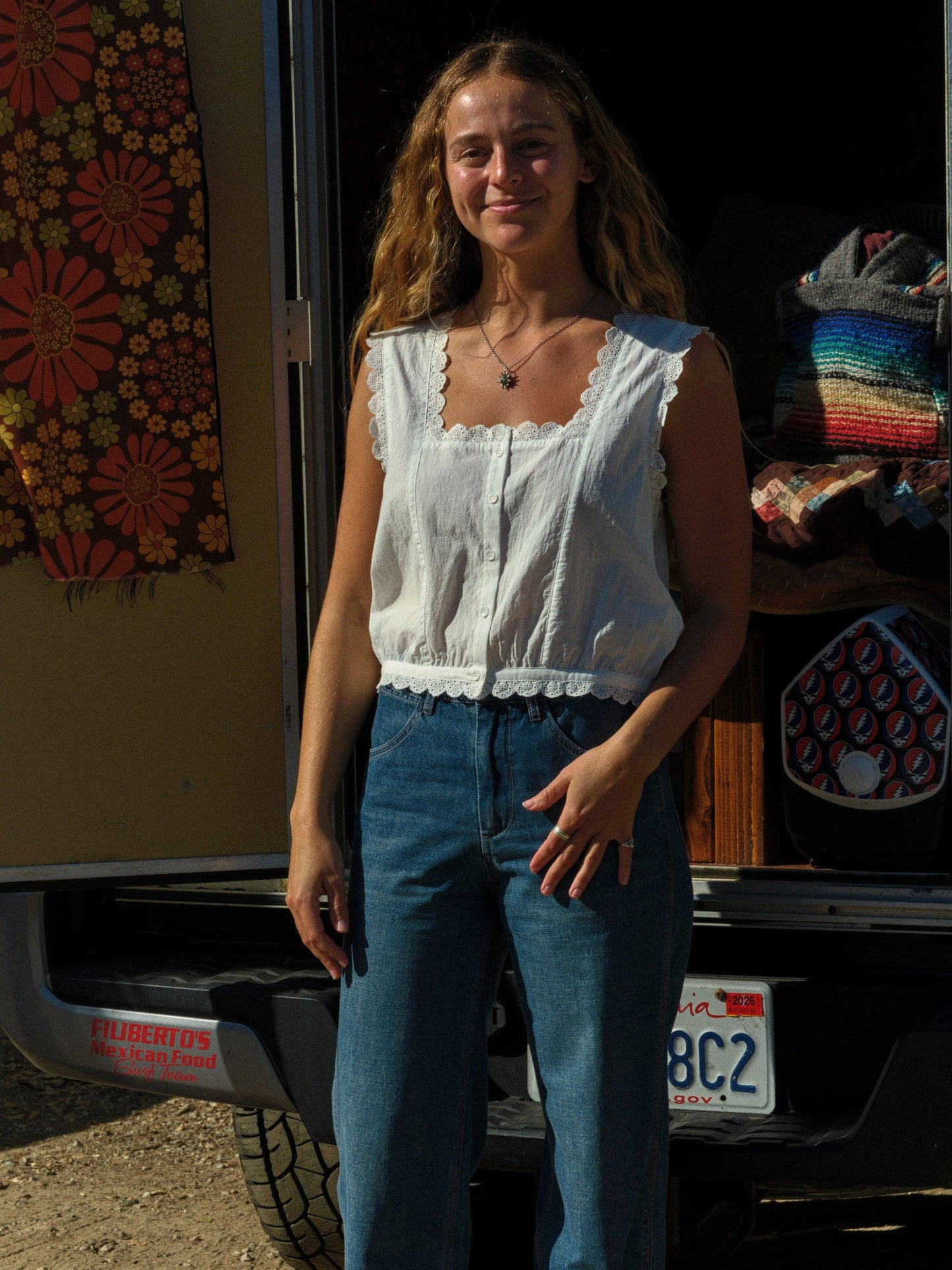 Woman wearing a cotton Helena top and blue jeans stands by an open van with layered blankets and a vibrant tapestry inside.