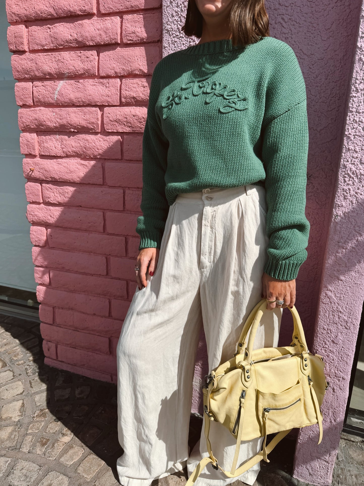 Woman in a cropped boat neck sweater holds pale yellow handbags against a pink brick wall. Sweater is knit; bags are leather.