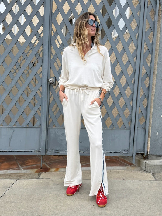 Woman in cream half zip sweatshirt and red sneakers stands with hands in pockets in front of a gray gate.