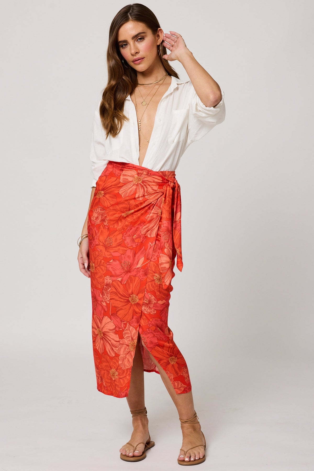Woman in a white shirt bodysuit and orange floral wrap skirt, barefoot, stands against a plain background.