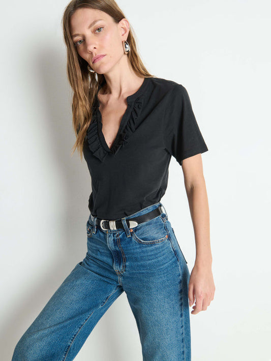 Woman in a black short-sleeve top, blue jeans, and earrings stands against a plain white background.