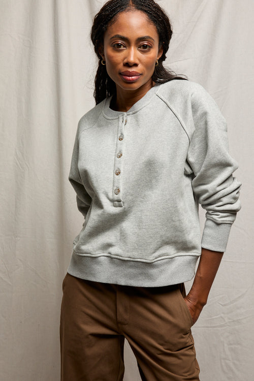 Woman in a medium heather grey sweatshirt with brown pants stands against a light background.
