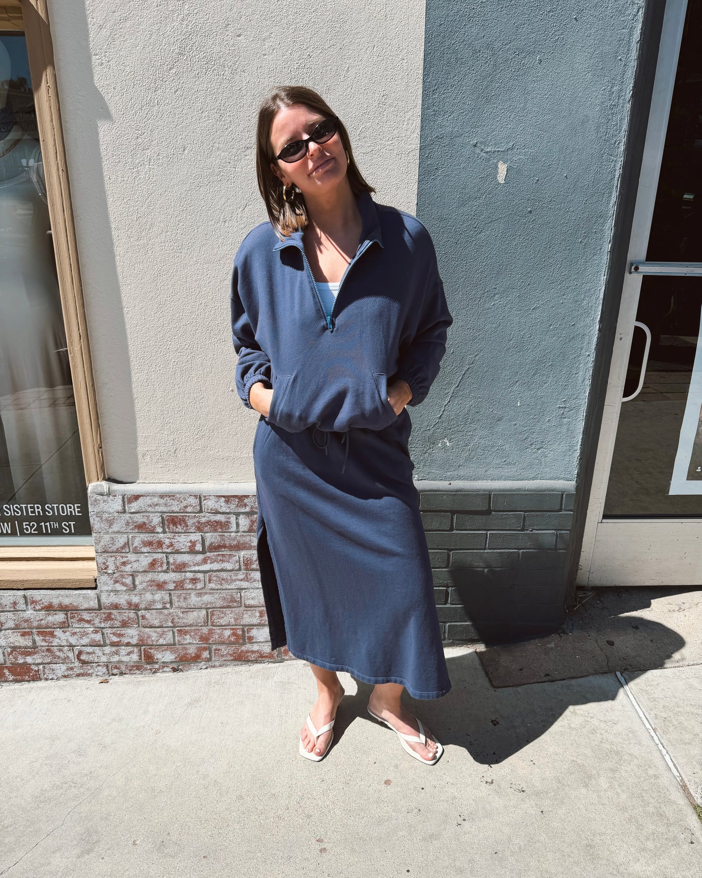 Woman wearing a navy half zip sweatshirt stands on a sunny sidewalk, smiling. Cotton blend, casual wear.
