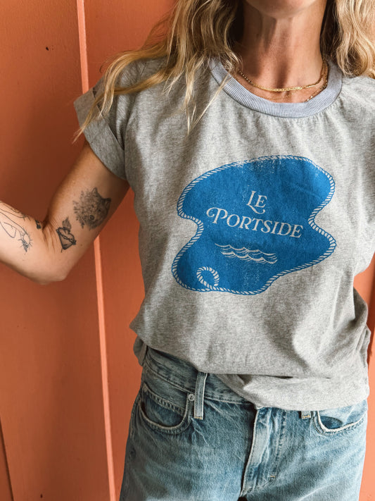 Person wearing a heather grey rolled-sleeve cotton tee and jeans stands by a coral wall, arm tattoos visible.