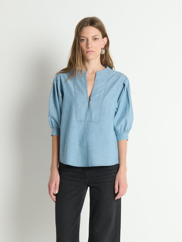Woman wears a chambray puff-sleeve Emmett top with black pants, standing against a plain white background.