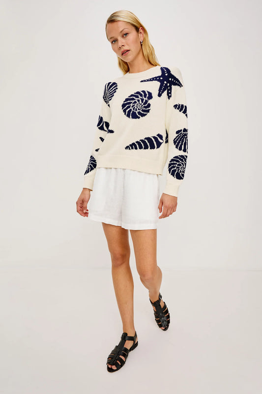 Woman in a light knit seashell-pattern sweater with white shorts and black sandals, standing against a plain background.