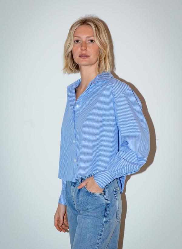 Woman in a blue and white pinstripe button-up shirt and blue jeans, standing against a plain white background.