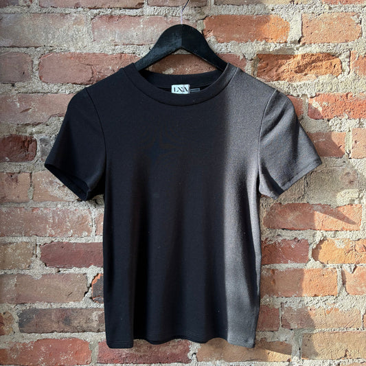 Black fitted crew neck tee, cotton material, displayed on a black hanger against a brick wall.