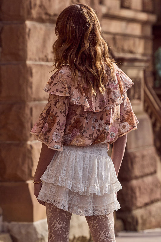 Woman in wavy hair wears a floral blouse and cream lace mini skirt near a stone building.