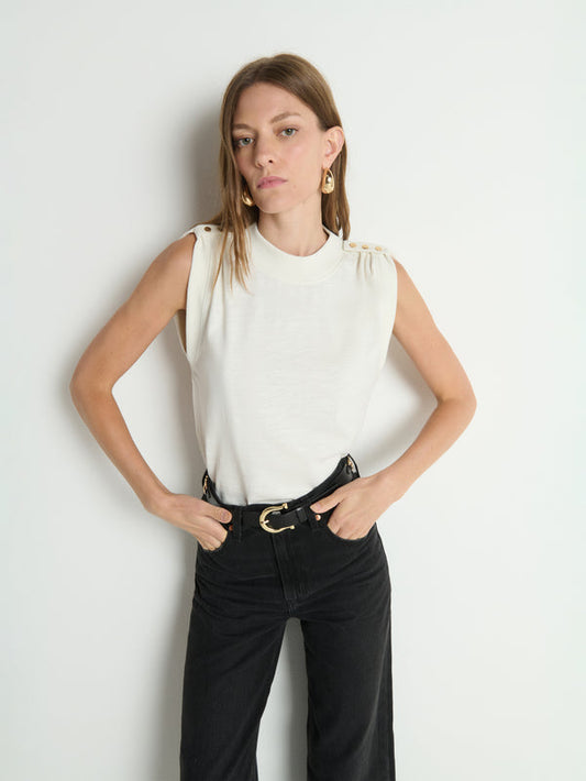 Woman in porcelain-white buttoned shoulder tank and black jeans stands with hands in pockets against a white wall.