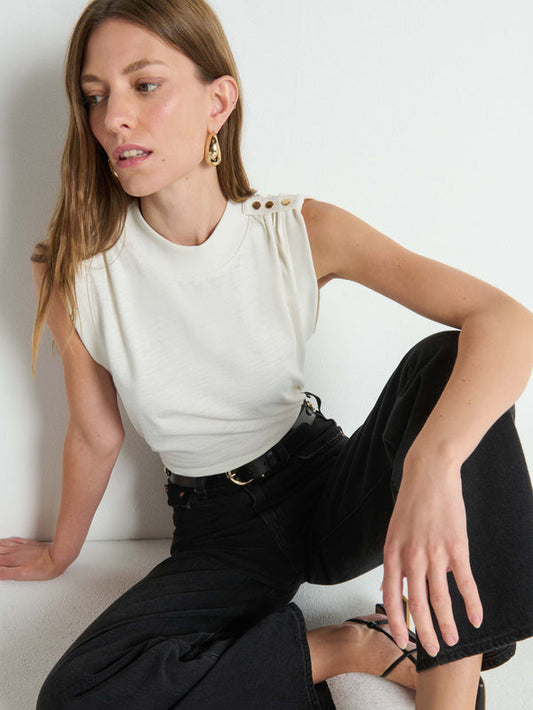 Woman with light brown hair wears an ivory buttoned-shoulder tank and black pants, standing beside a white wall.