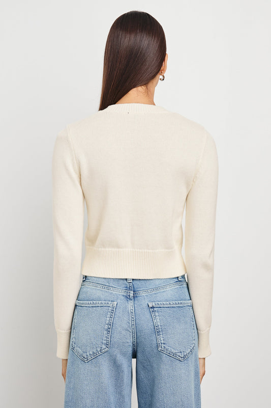 Back view of a woman wearing an ivory knit cardigan and blue jeans, standing against a plain white background.