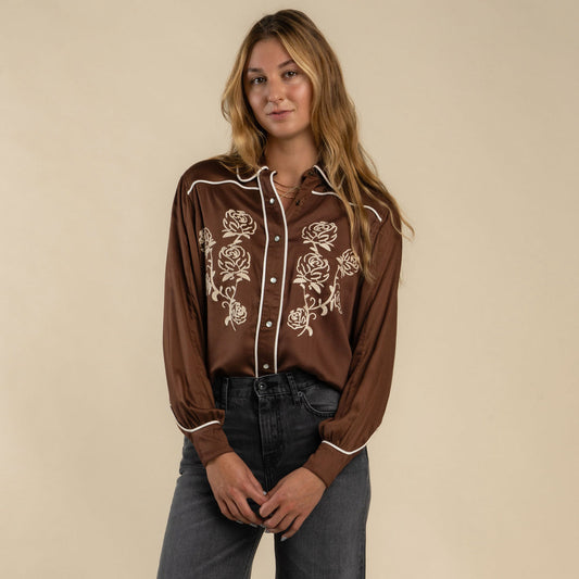 Woman wearing a mocha embroidered long sleeve shirt and dark gray jeans, suitable for casual or outdoor wear.
