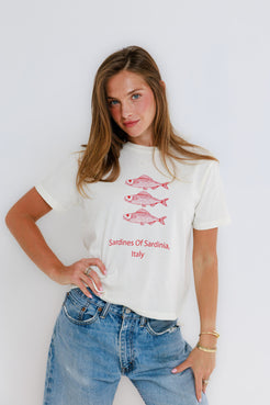 Woman models white cotton tee with three pink fish, standing against a white wall. Suitable for casual wear.