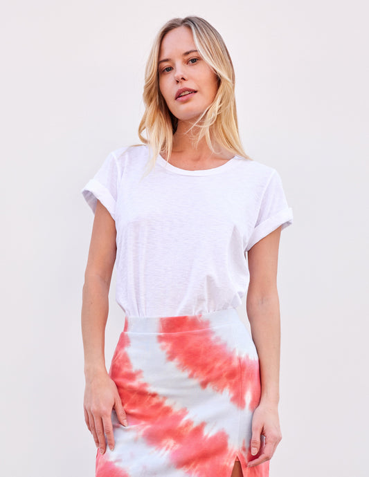Woman wearing optic white rolled sleeve tee and pink tie-dye skirt, standing in front of a plain light background.