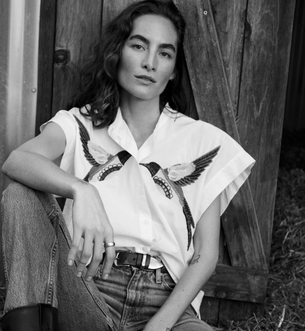 Woman with wavy hair by a wooden door wearing a white cotton blouse and jeans; black and white photo.