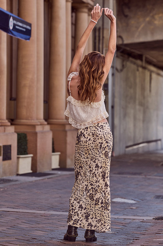 Woman in a lace top and leopard print maxi skirt stretches her arms overhead while standing on the street.