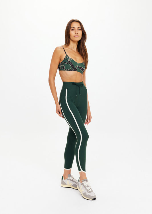 Woman in studio wears green recycled sports bra and leggings with gray sneakers, suitable for active or casual wear.