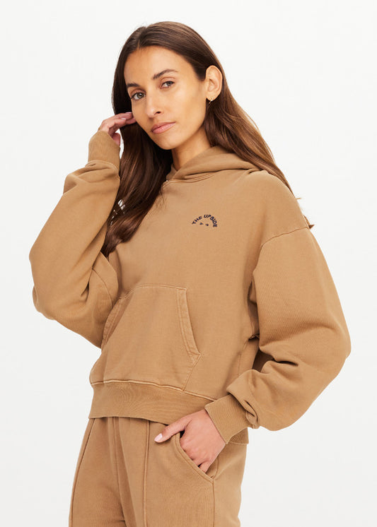Woman wearing a chai-colored Delphi hoodie with one hand in her hair, shown against a plain white background.