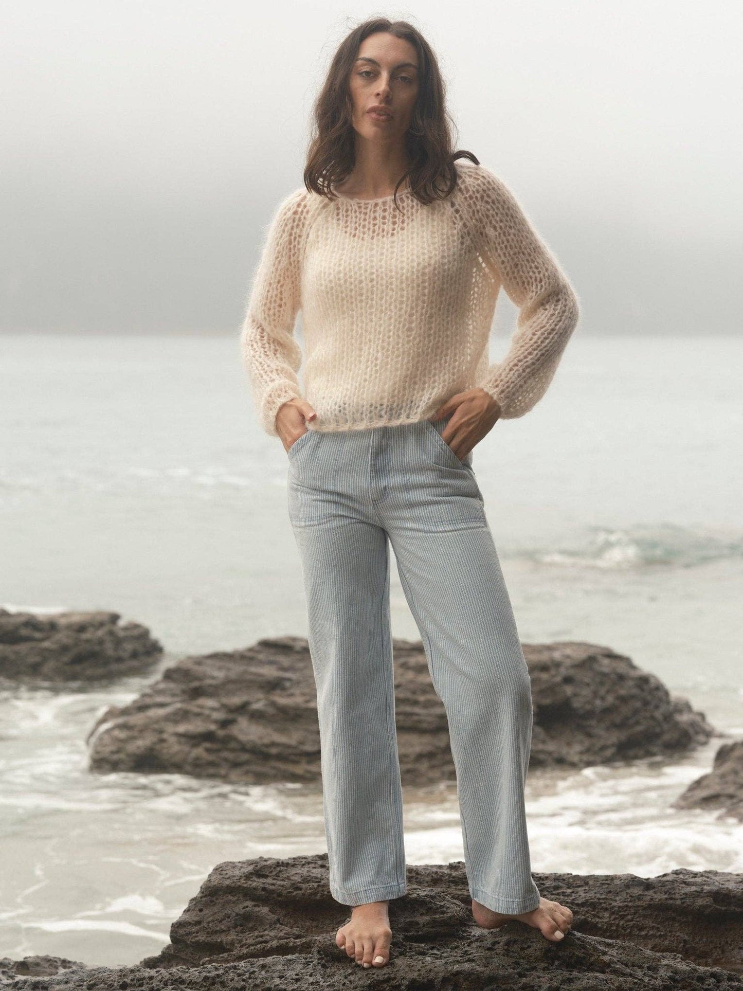 Woman wears cream knit sweater and light jeans, standing barefoot on coastal rocks in ocean mist.