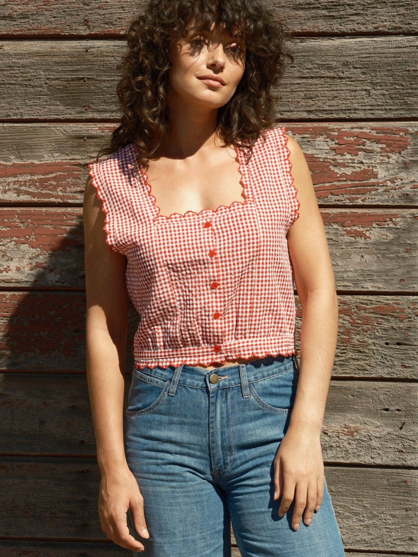 Sleeveless gingham square-neck top with blue jeans, worn by a rustic wooden wall.
