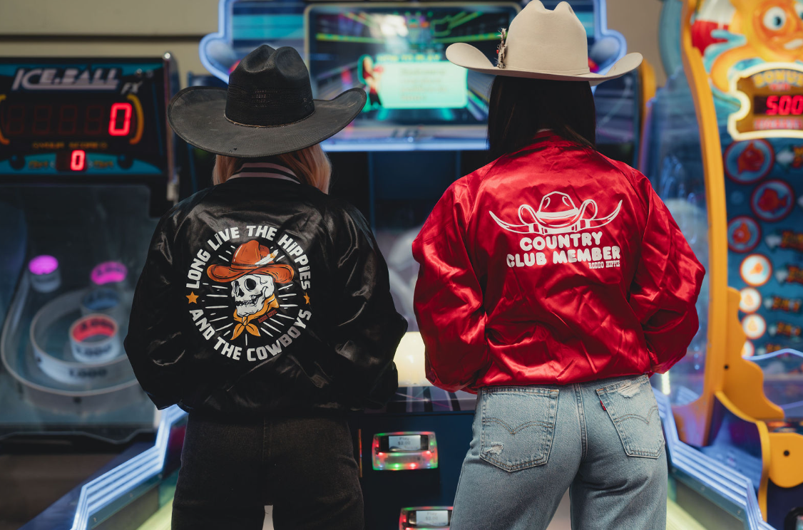 Two people in satin COUNTRY CLUB MEMBER jackets and cowboy hats play arcade games, backs to camera.
