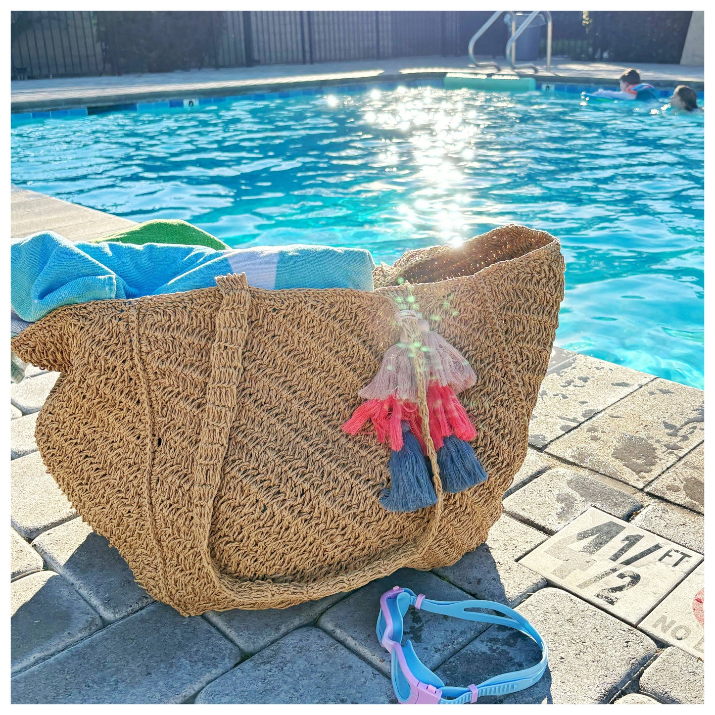 A woven canvas tote in neutral tones rests beside a sunlit pool, placed on the deck near people swimming.