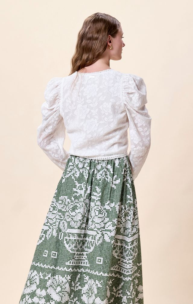 Woman in white cotton blouse and green patterned skirt, seen from behind, wavy hair visible.