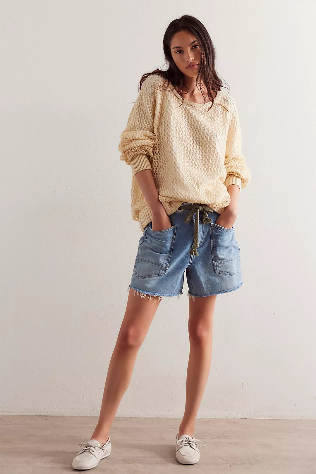 Person wearing loose blue denim shorts, cream knit sweater, and white shoes standing near a light-colored wall.