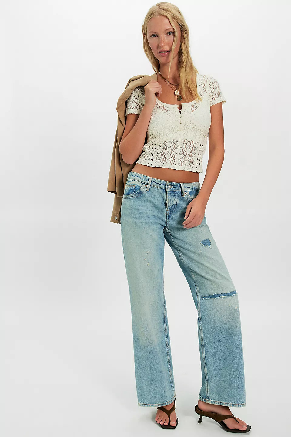 Woman wearing a white lace tee, light-wash wide-leg jeans, sandals, and carrying a tan jacket over her shoulder.