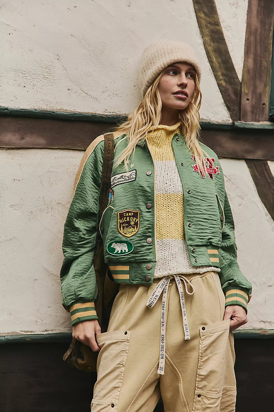 Woman in a vintage green souvenir jacket and beanie stands by a rustic wooden wall.