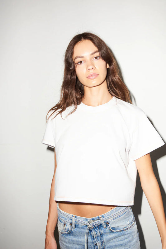 Woman in a white cotton tee and blue jeans stands against a plain wall, facing the camera.