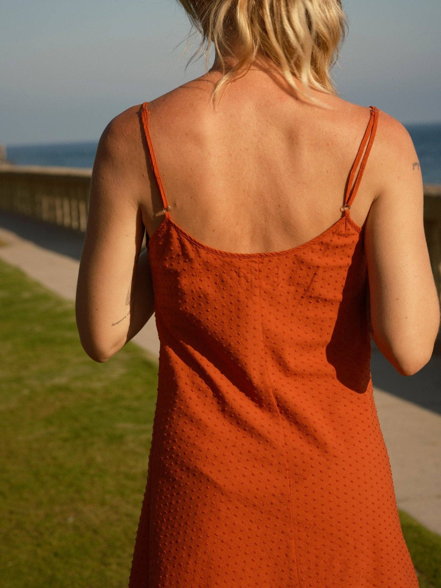 Blonde person wearing a light-colored dobby slip dress stands outside near grass and a stone wall, back to camera.