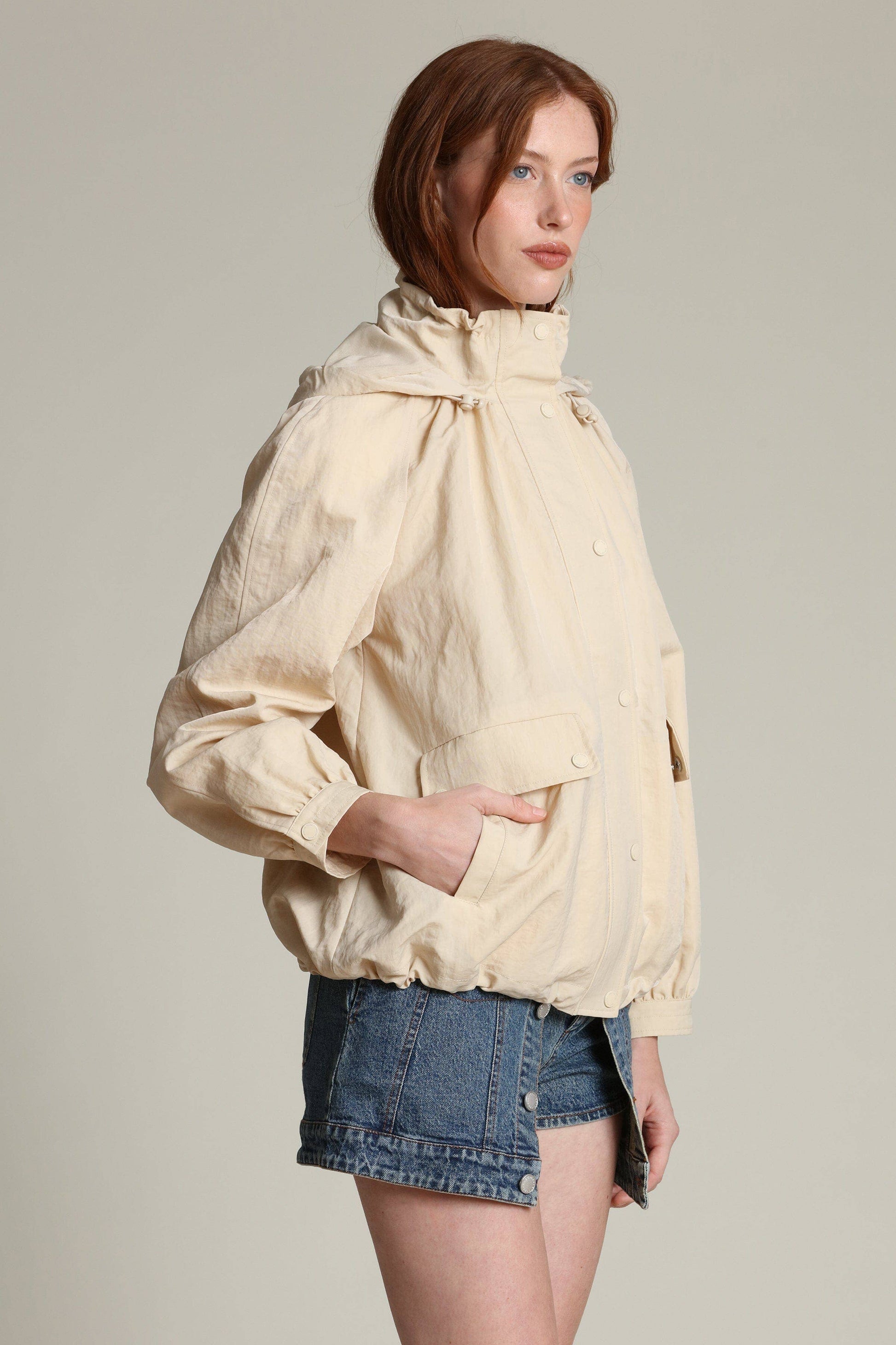 Woman in a light windbreaker with balloon sleeves and denim shorts, posed sideways against a plain backdrop.