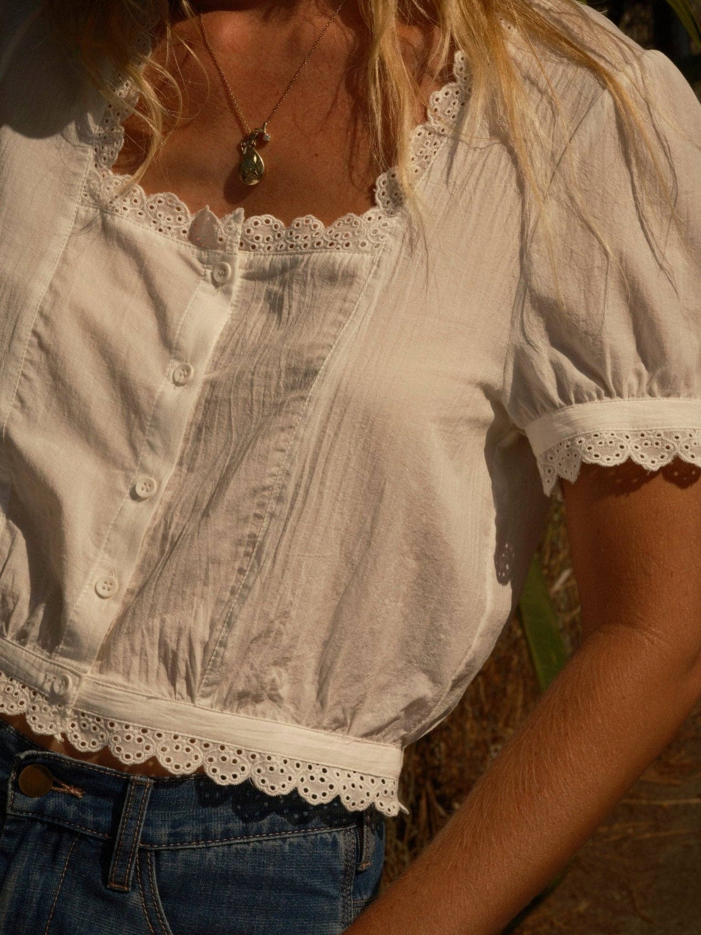 Woman outdoors in white scalloped eyelet top, blue jeans, and necklace, standing in sunlight.