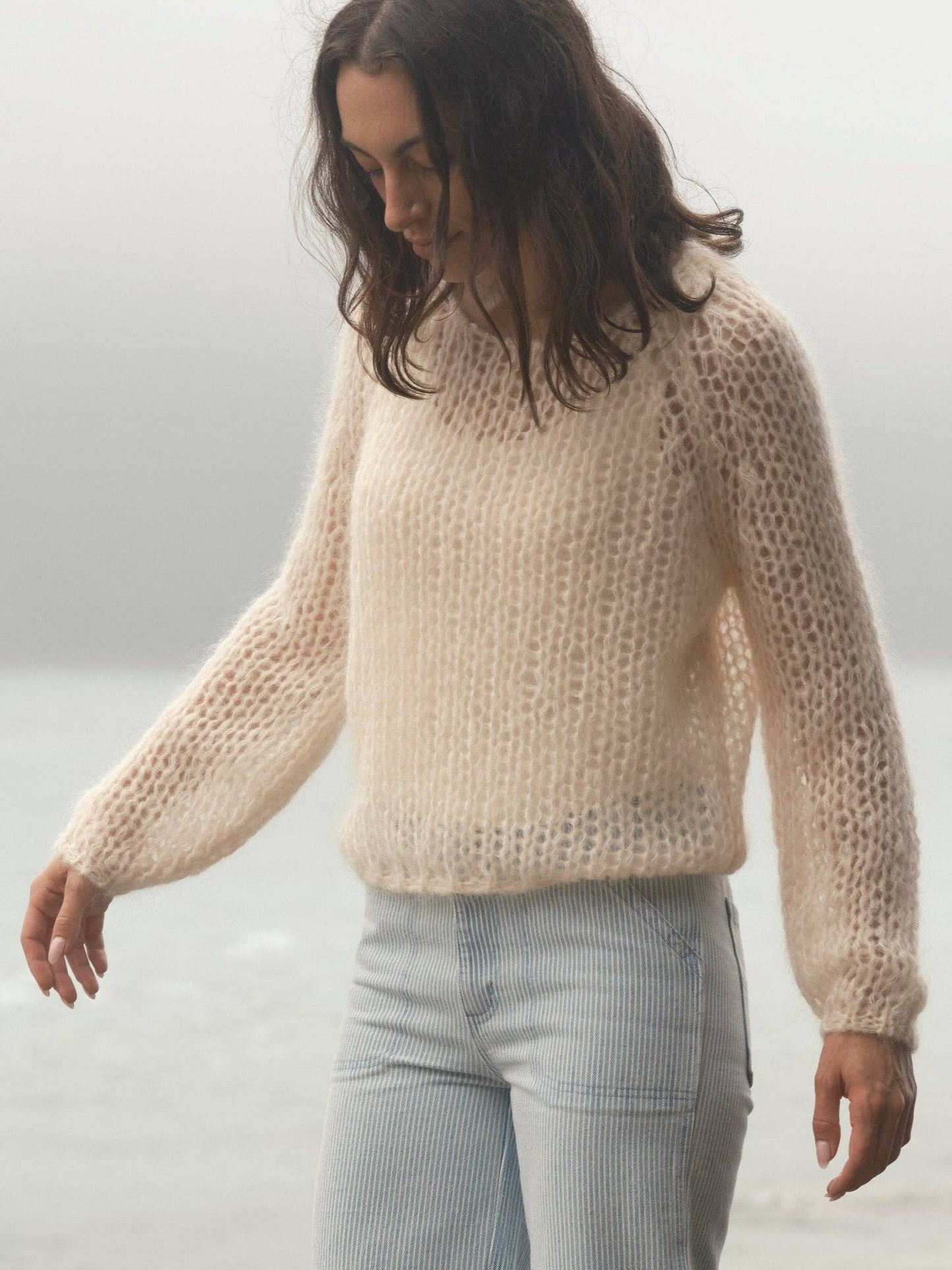 Woman wearing a sheer cream sweater and light pants stands by a foggy waterside, looking down.