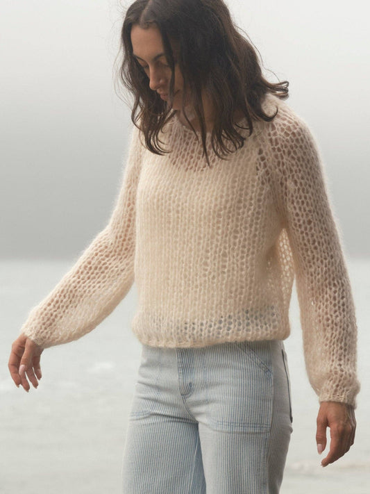 Woman wearing a sheer cream sweater and light pants stands by a foggy waterside, looking down.