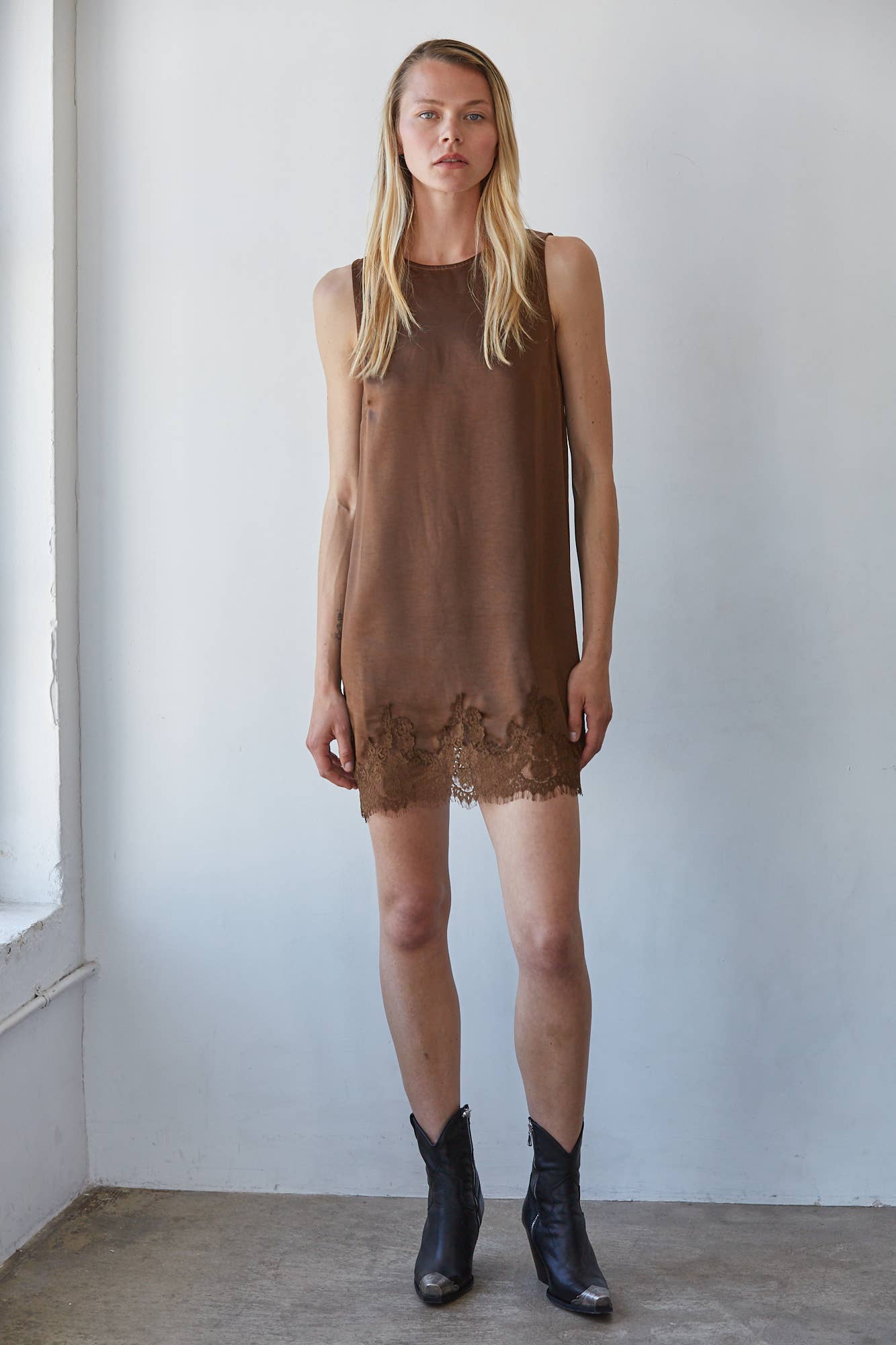 Brown sleeveless mini dress with lace hem, worn by a woman standing against a white wall; ideal for warm weather occasions.