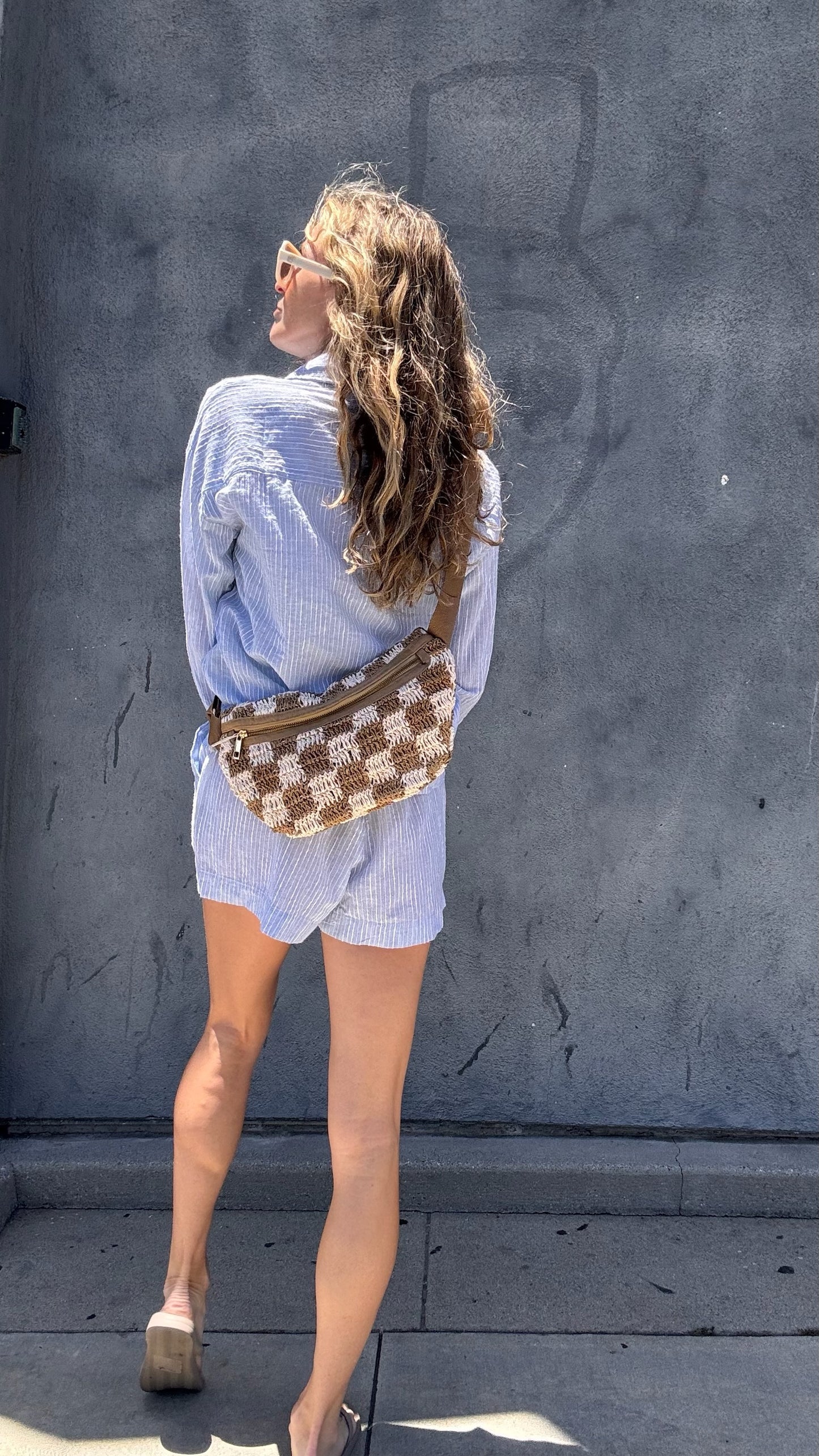 Woman with wavy hair in light blue shorts and sandals, holding a woven crossbody bag by a gray wall.