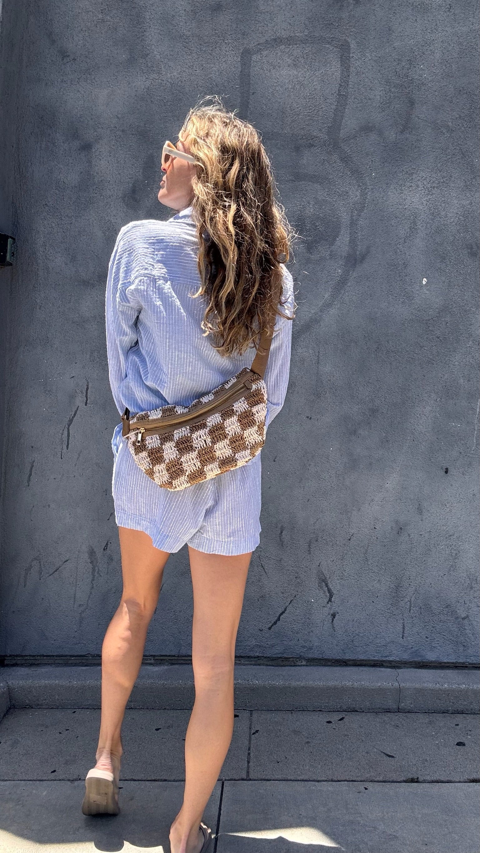Woman with wavy hair in light blue shorts and sandals, holding a woven crossbody bag by a gray wall.