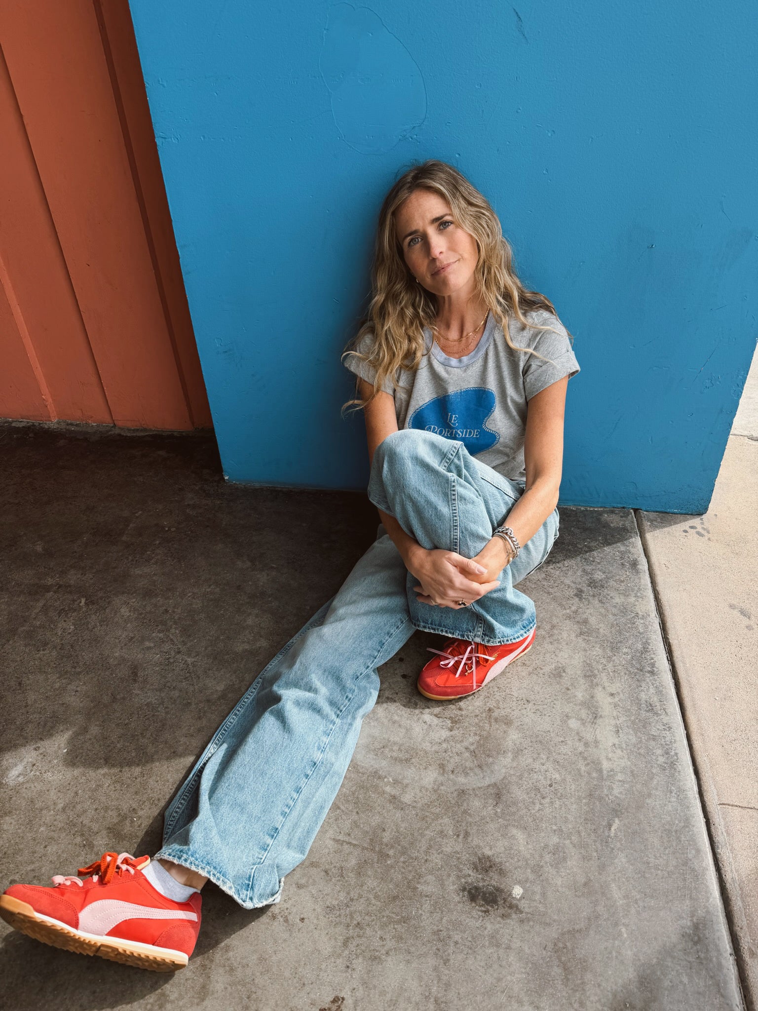 Woman wearing a heather grey rolled sleeve tee sits by a blue wall, gazing thoughtfully at the camera.