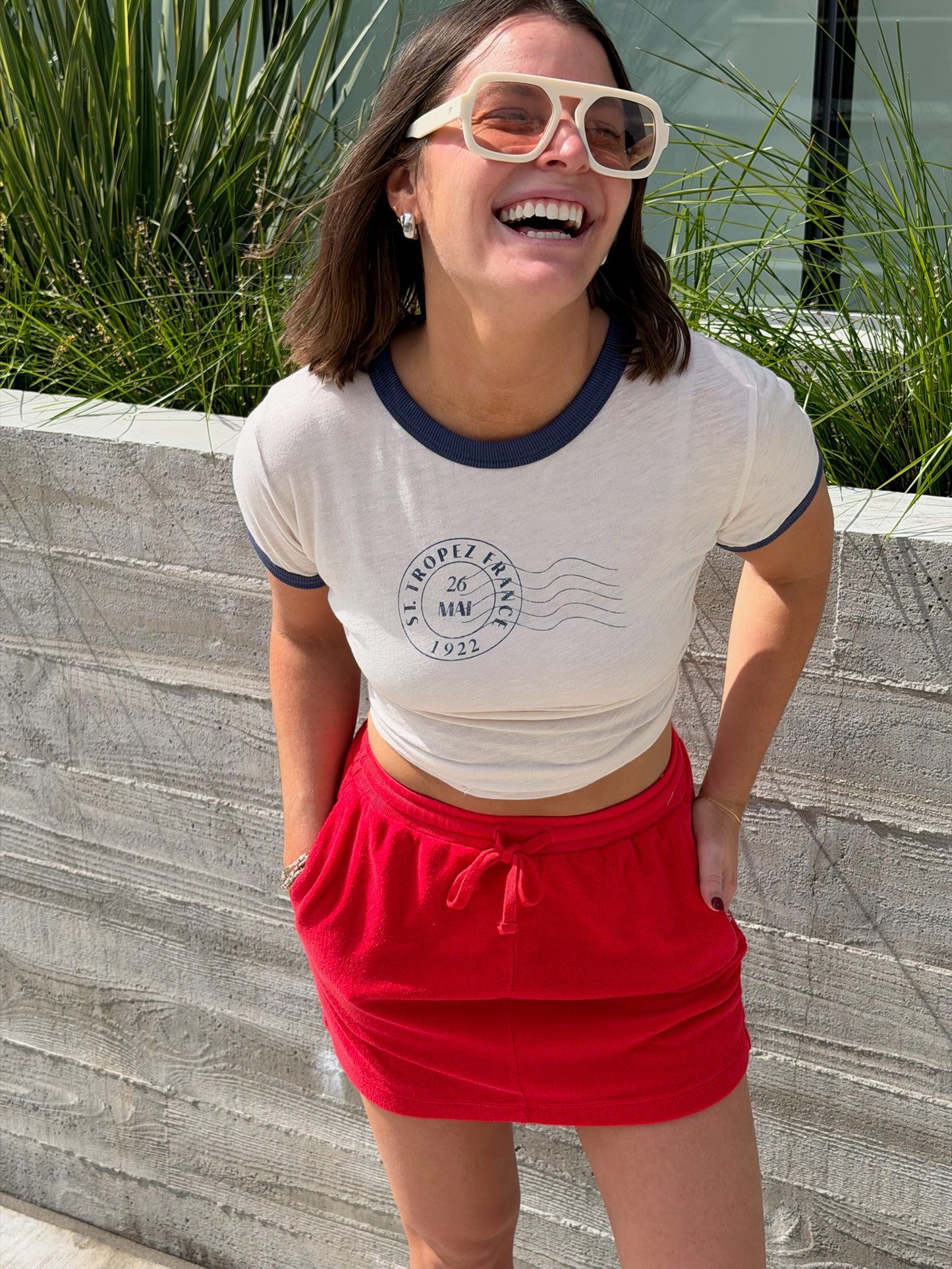 Woman outdoors in a vermillion terry mini skirt, cream crop top, white sunglasses, hands in skirt pockets.