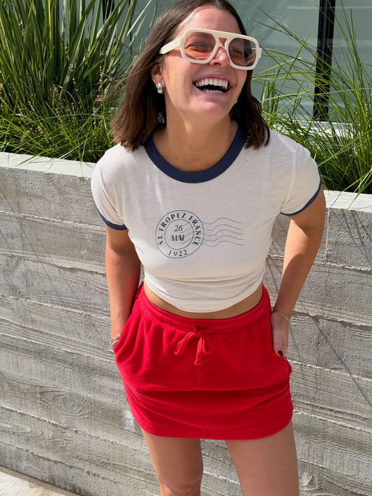 Woman outdoors in a vermillion terry mini skirt, cream crop top, white sunglasses, hands in skirt pockets.