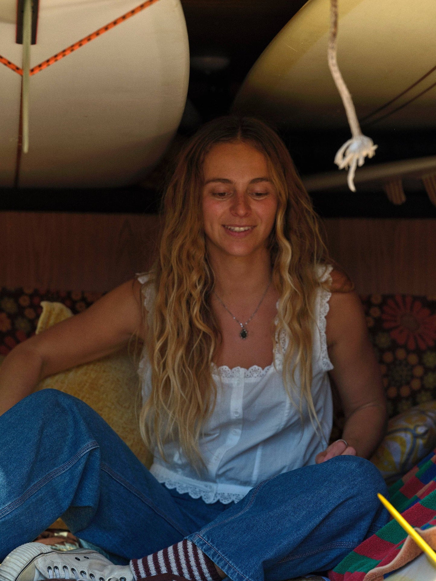 Woman with long wavy hair wears a blue cotton top, sitting in a van with surfboards and pillows in the background.
